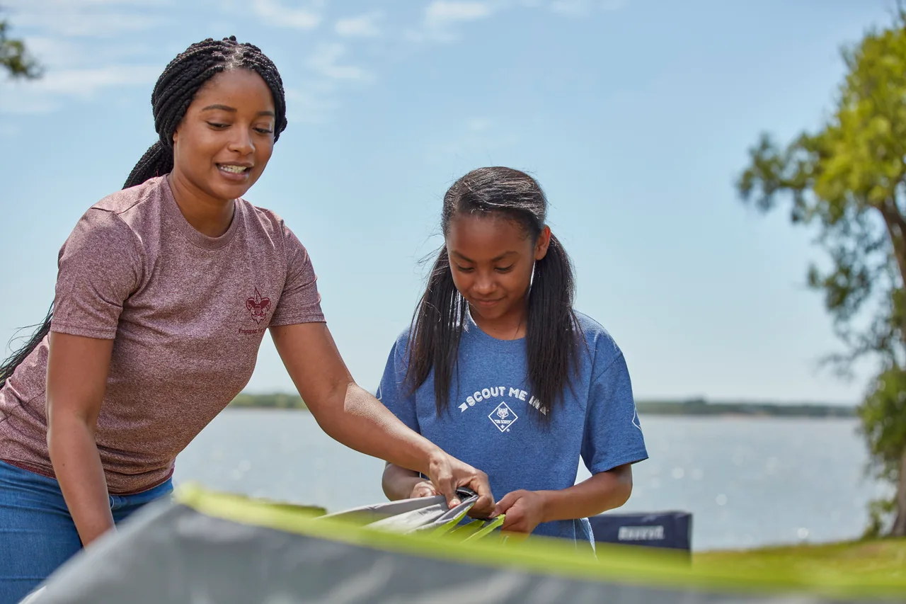 A parent and Scout work together to set up a tent beside a lake