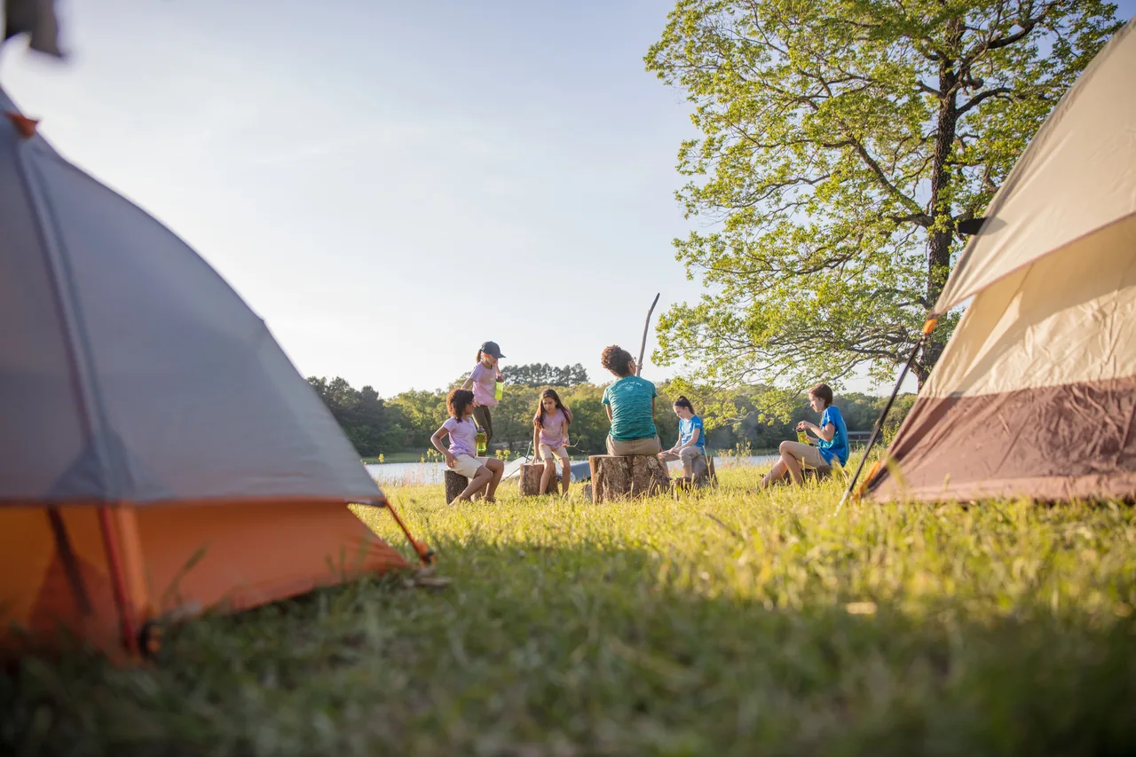 Girls in Scouts BSA gather at a campsite between tents near a lake